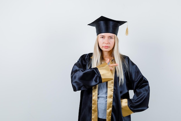 Chica rubia en toga y gorra de graduación apuntando hacia la derecha con el dedo índice y luciendo linda