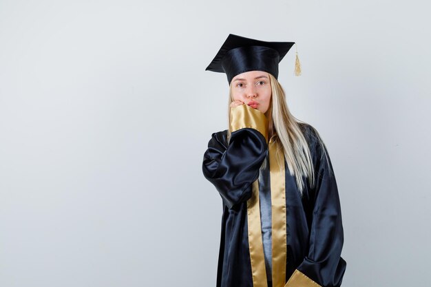 Chica rubia en toga y gorra de graduación enviando besos y poniendo la mano cerca de la boca y luciendo linda