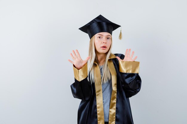 Chica rubia en toga y gorra de graduación levantando las manos en posición de rendición y mirando asustada