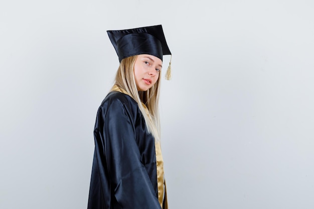 Chica rubia en toga y gorra de graduación mirando por encima del hombro y posando a la cámara y luciendo linda