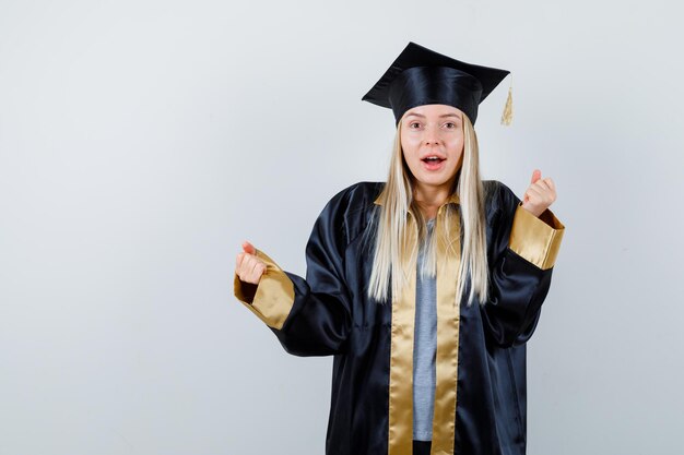 Chica rubia en toga y gorra de graduación mostrando gesto de ganador y mirando feliz