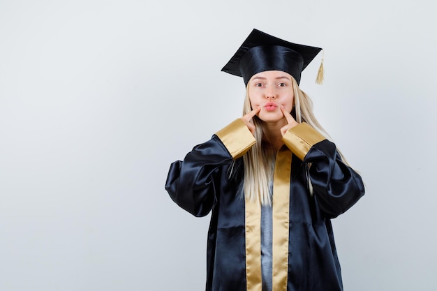 Foto gratuita chica rubia en toga y gorra de graduación poniendo los dedos índices cerca de la boca y luciendo linda