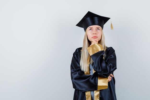 Chica rubia en uniforme graduado apoyando la barbilla en la mano y mirando pensativo