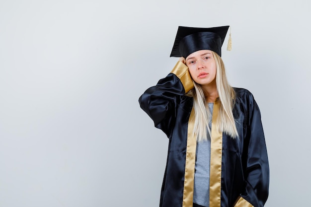 Chica rubia en uniforme graduado sosteniendo la mano en la cabeza y mirando triste