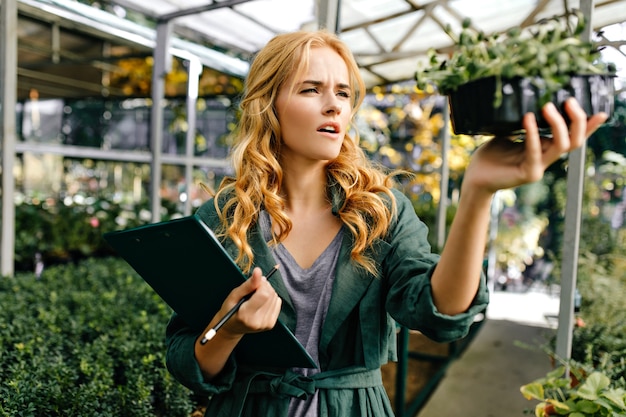 Chica rubia, vestida con camiseta gris y túnica verde oscuro, sostiene una maceta con una planta con hojas pequeñas.