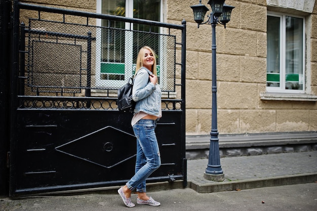 Chica rubia vestida con chaqueta de jeans con mochila posada en las calles de la ciudad