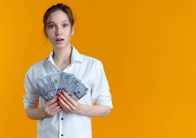 Chica rusa rubia sorprendida joven tiene dinero aislado en el espacio naranja con espacio de copia