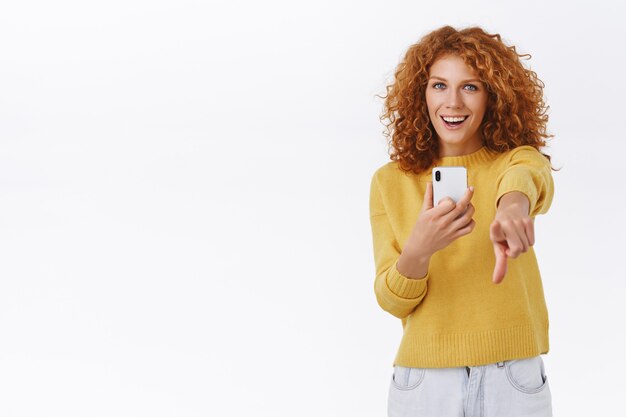 Chica satisfecha con tu posando como fotografiando en smartphone. Atractiva mujer rizada pelirroja emotiva sostenga el teléfono móvil, apuntando la cámara con el dedo índice, sonriendo con alegría, pared blanca