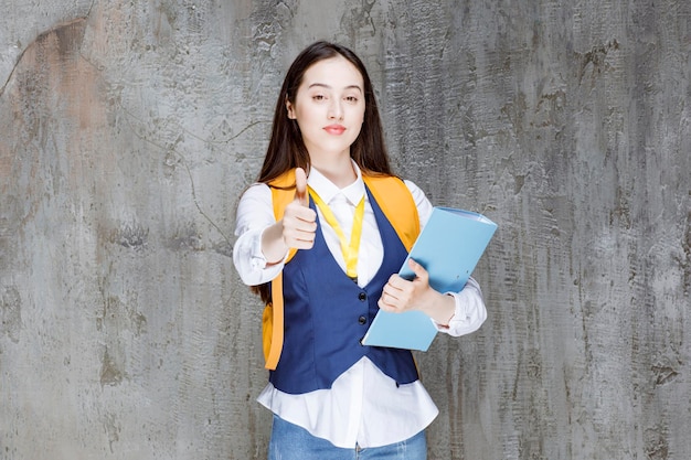 Chica de secundaria con mochila amarilla y carpeta dando pulgares hacia arriba. foto de alta calidad