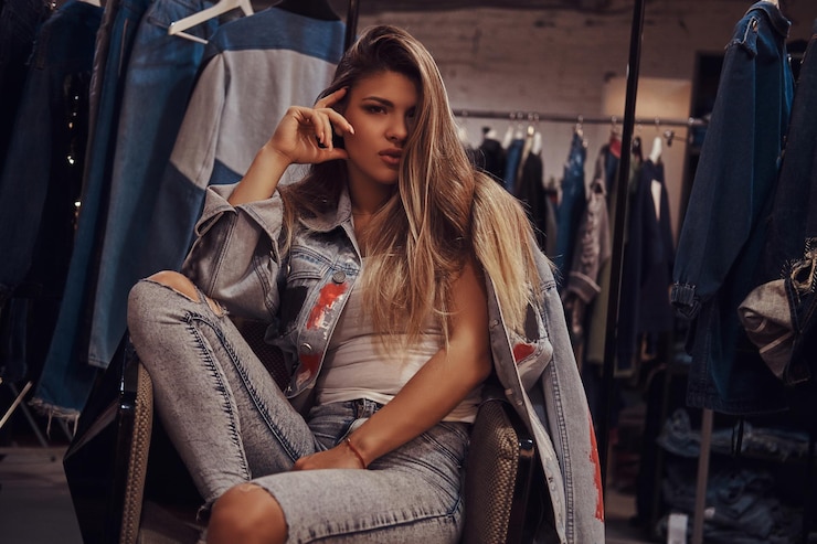 Foto gratuito chica seductora con jeans desgastados y chaqueta de mezclilla mirando una cámara mientras se sienta en una silla en el probador de una tienda de ropa.