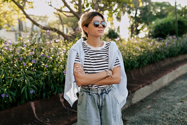 Chica segura en elegante traje de verano con gafas de sol con una sonrisa encantadora descansando al aire libre día soleado Retrato de mujer joven alegre esperando amigos en el jardín de la ciudad