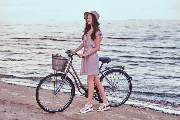 Chica sensual feliz con sombrero vestido camina con su bicicleta en la playa contra un increíble fondo costero.