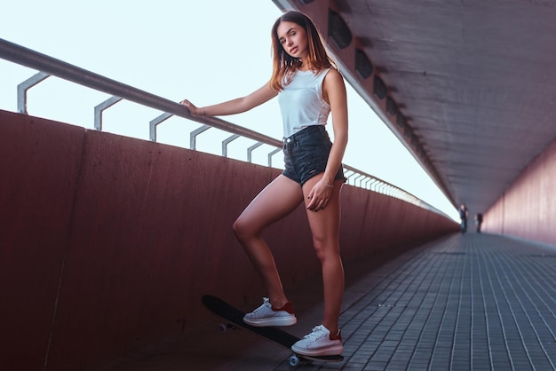 Chica sensual joven vestida con pantalones cortos y camiseta de pie en una patineta mientras se apoya en la barandilla en una acera bajo el puente.