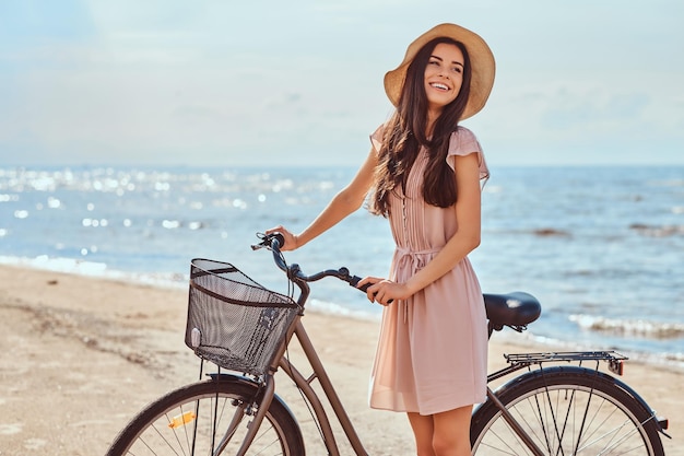 Chica sensual riendo con cabello castaño vestido con vestido y sombrero posando con bicicleta en la playa en un día soleado.