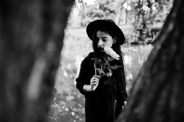 Chica sensual toda en labios rojos negros y sombrero Goth mujer dramática sostiene flor de crisantemo blanco a mano contra el lago silencioso Retrato en blanco y negro