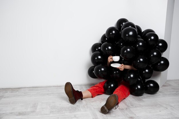 Chica sentada en globos negros sosteniendo una taza sobre una pared blanca