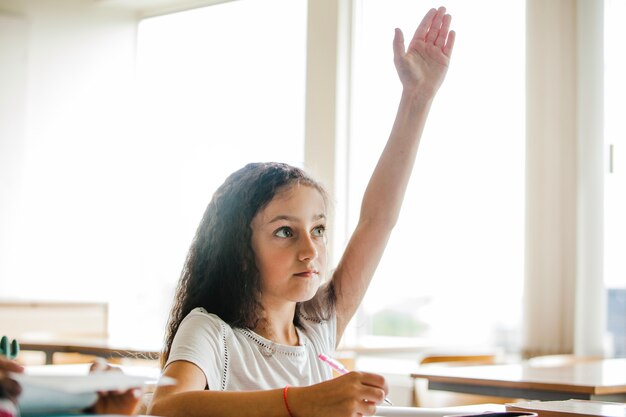 Chica sentada en la mesa de la escuela levantando la mano