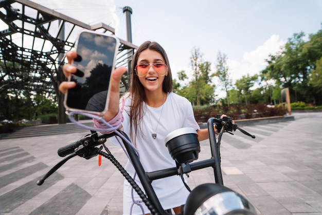 Chica sentada en un scooter y mostrando el teléfono a la cámara