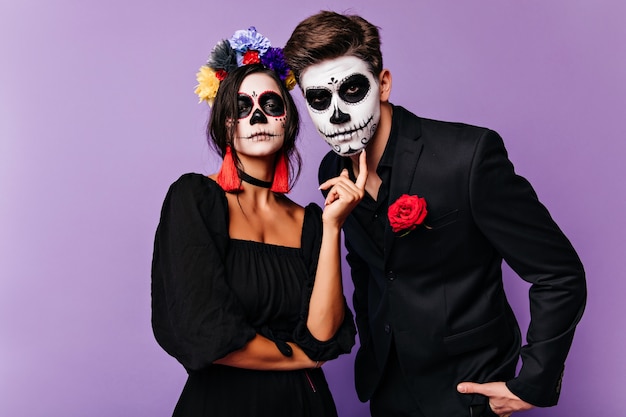 Chica seria en traje de mascarada posando sobre fondo morado. Pareja divertida en trajes de carnaval mirando a la cámara.