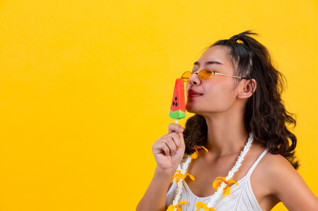 Chica sexy está oliendo el helado de sandía mientras usa gafas en clima caluroso de verano en la pared amarilla