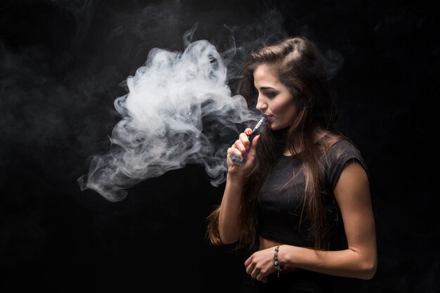 Chica sexy en un vestido negro fumar cigarrillo electrónico en la pared oscura