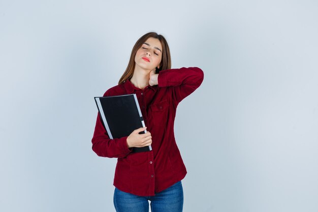 chica siente dolor de cuello mientras sostiene la carpeta en camisa burdeos y se ve cansada.