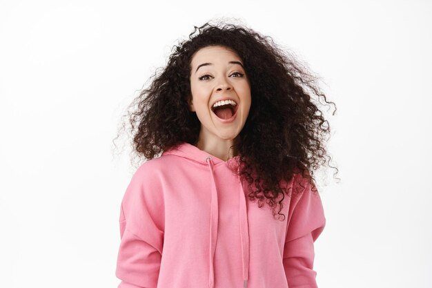 Chica sincera feliz con cabello rizado oscuro, riendo, sonriendo y mirando asombrada a la cámara, divirtiéndose, mostrando emoción, de pie sobre fondo blanco