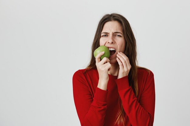 Chica sintiendo dolor de muelas y haciendo muecas de dolor mientras muerde una manzana verde