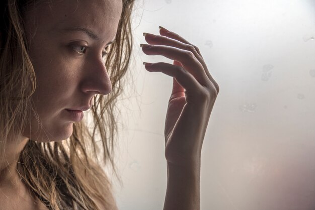 Chica solitaria cerca de la ventana pensando en algo. Mujer triste mirando la lluvia caer a través de una ventana en casa