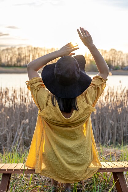 Una chica con sombrero y estilo casual se sienta en un banco cerca del lago al atardecer