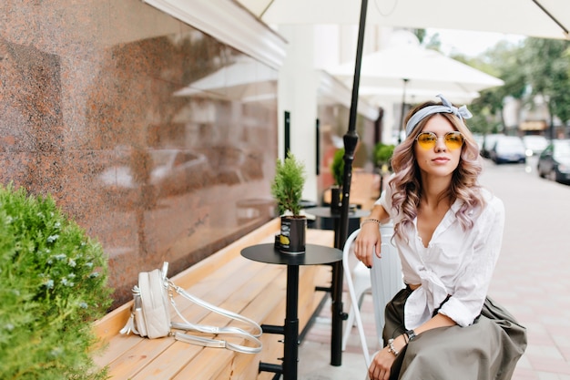 Chica soñadora en camisa blanca con mochila pequeña esperando novio en café al aire libre