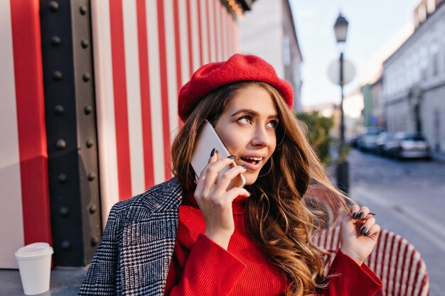 Chica soñadora en linda boina roja juega con el pelo oscuro mientras habla por teléfono