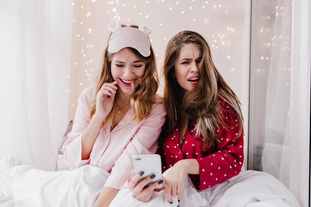 Chica soñadora en máscara de dormir riendo mientras está sentado en la cama. Infeliz dama de cabello castaño leyendo un mensaje de teléfono en el dormitorio.