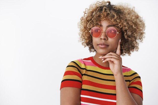 Chica soñadora mirando a otro lado como pensando. Encantadora mujer afroamericana con estilo en traje de moda de los años 90 con gafas de sol rojas frescas mirando en la esquina superior izquierda pensativo y relajado con una leve sonrisa