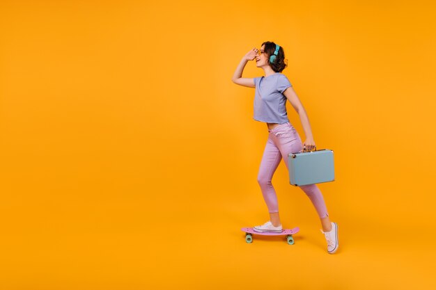 Chica soñadora en pantalones rosas de pie en patineta y escuchando música. Modelo femenino rizado inspirado en auriculares posando con valija azul.