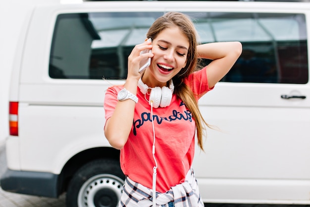 Chica sonriente alegre en camisa rosa de moda disfrutando de la música favorita de pie cerca del coche blanco