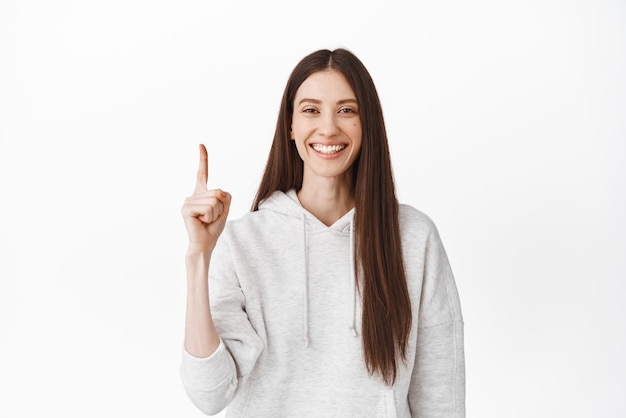 Chica sonriente alegre con una sonrisa perfecta y cara limpia apuntando con el dedo hacia arriba en el logotipo superior que muestra un cartel publicitario encima de pie en un fondo blanco casual con capucha