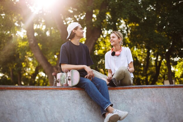 Chica sonriente con auriculares y chico joven con monopatín alegremente pasando tiempo juntos en el skatepark moderno