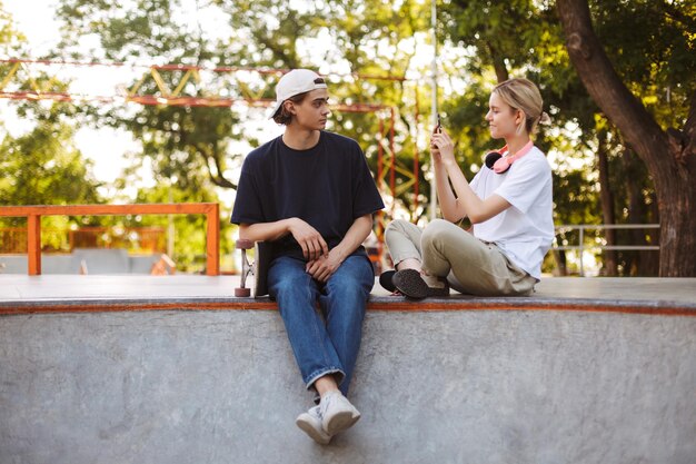 Chica sonriente con auriculares tomando fotos de un joven con patineta en el celular pasando tiempo juntos en el skatepark moderno