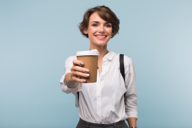 Chica sonriente con cabello corto oscuro en camisa blanca mostrando felizmente en la cámara una taza de café para repasar el fondo azul aislado