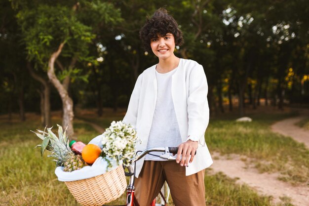 Chica sonriente con camisa blanca en bicicleta clásica y cesta llena de frutas y flores silvestres mirando alegremente a la cámara pasando tiempo en el parque