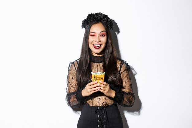 Chica sonriente descarada en traje de bruja, celebrando halloween, haciendo truco o trato con un vestido gótico, mostrando la lengua y sosteniendo dulces