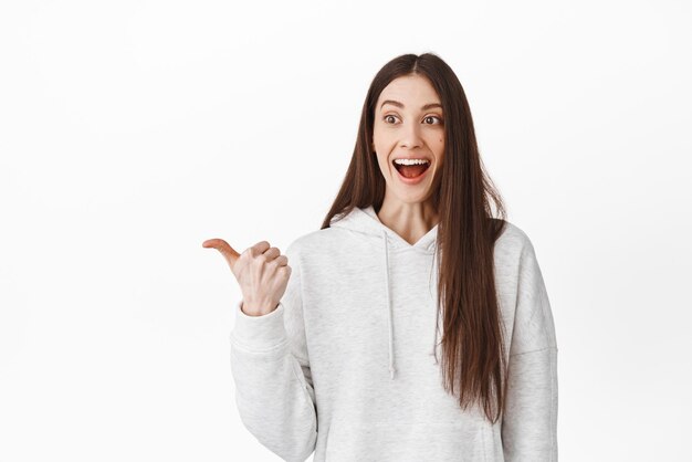 Una chica sonriente divertida dice wow sonriendo y mirando asombrada al espacio de la copia del lado izquierdo que muestra un impresionante logotipo de texto promocional o pancarta a un lado de pie sobre fondo blanco