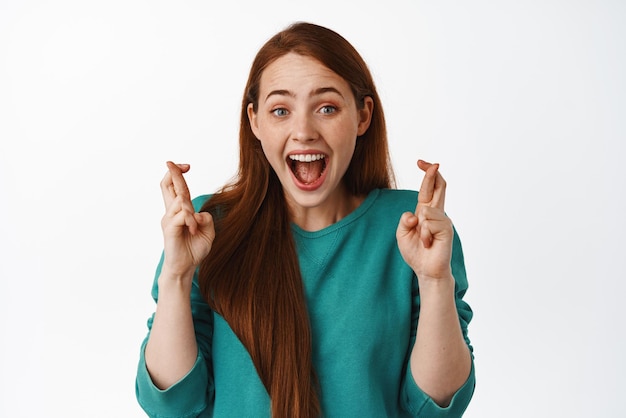 Chica sonriente emocionada esperando un resultado positivo con la cara feliz optimista cruza los dedos y pide deseos deseando ganar de pie sobre fondo blanco