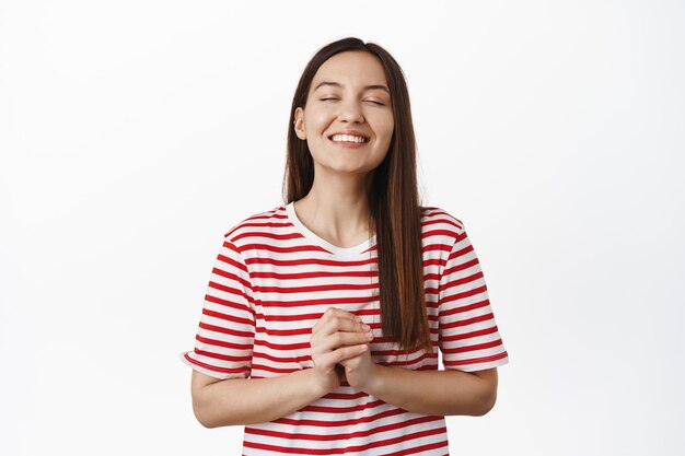 Una chica sonriente y esperanzada cierra los ojos, tomándose de la mano, rezando o pidiendo un deseo, esperando que suceda algo, soñando despierto, imaginándose en una burbuja sobre la cabeza, fondo blanco