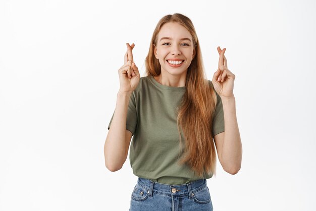 Chica sonriente esperanzada cruza los dedos para tener buena suerte y sonríe feliz esperando buenas noticias rezando anticipando ganar de pie sobre fondo blanco