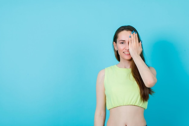 Chica sonriente está cubriendo la mitad de la cara con la mano sobre fondo azul.