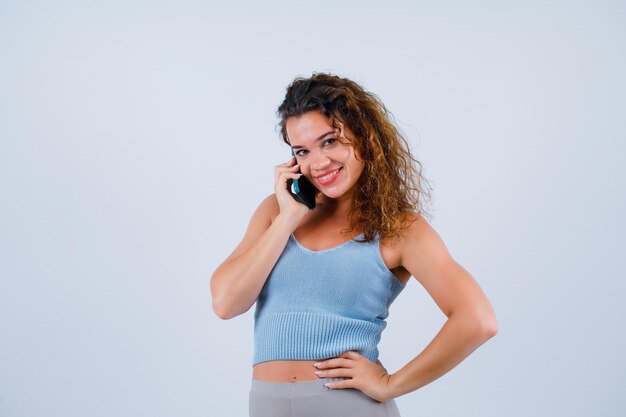 Chica sonriente está hablando por teléfono poniendo la mano en la cintura sobre fondo blanco.