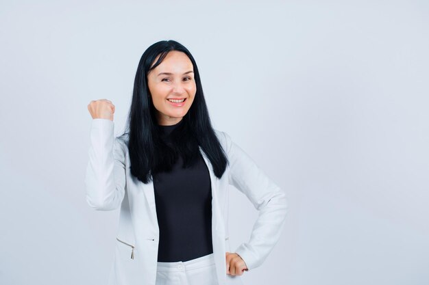 La chica sonriente está levantando el puño y poniendo la otra mano en la cintura sobre fondo blanco.