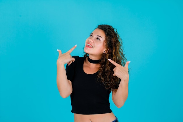 La chica sonriente está mirando hacia arriba apuntándose a sí misma con los dedos índices sobre fondo azul.